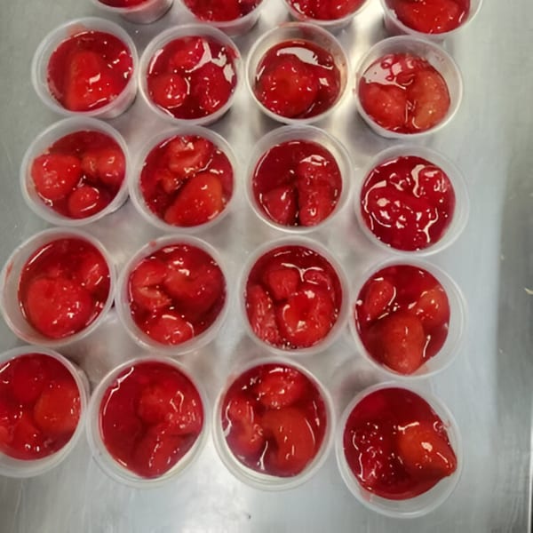 Rows of small plastic cups filled with bright red strawberry topping on a metal surface.