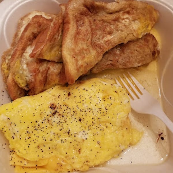 Scrambled eggs with black pepper and French toast in a white takeout container with a plastic fork.