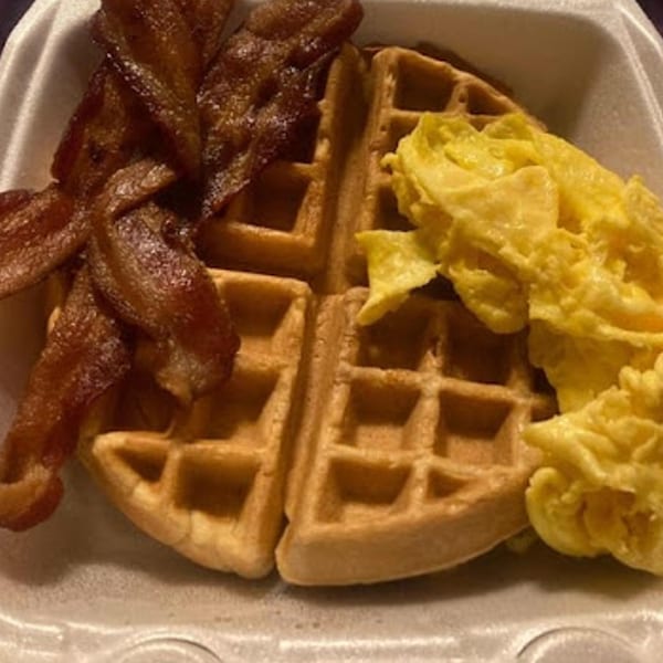 A breakfast of crispy bacon, fluffy scrambled eggs, and two golden waffles in a takeout container.