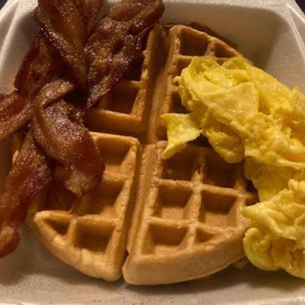 A breakfast of crispy bacon, fluffy scrambled eggs, and a golden waffle in a takeout container.